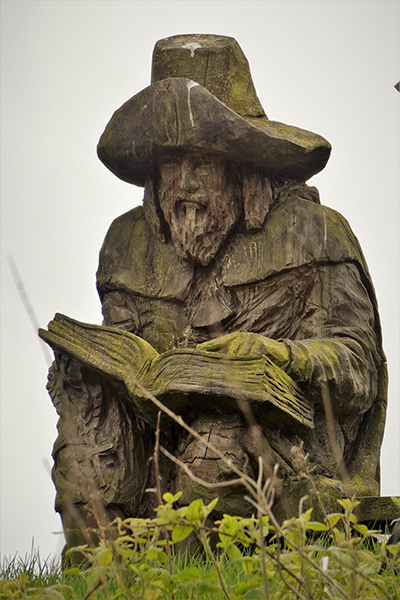 Cerflun pren o ddyn barfog mewn het lydan yn darllen llyfr, wedi'i orchuddio'n rhannol â mwsogl, wedi'i osod yn erbyn awyr gymylog gyda glaswellt a dail yn y blaendir